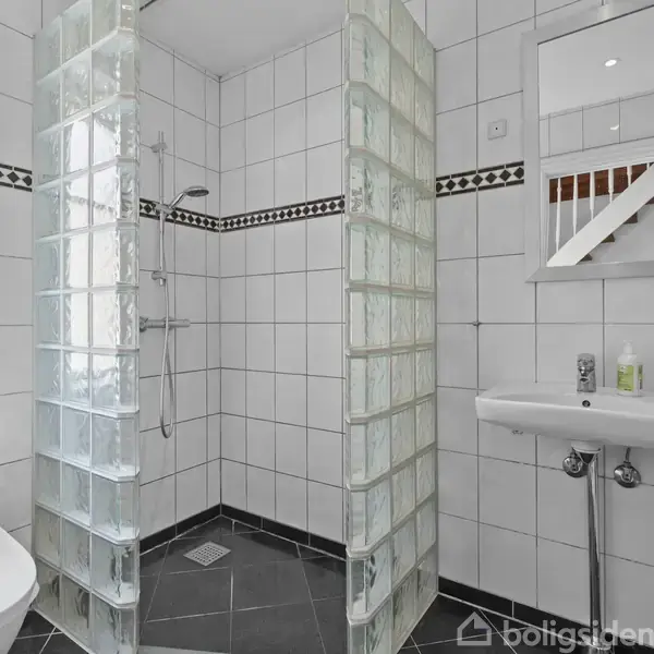 Et badeværelse viser en bruseniche af glassten med et brusehoved; ved siden af ses en vask med et spejl over. Væggene er flisebelagte, og der er en toiletkumme ved vinduet.