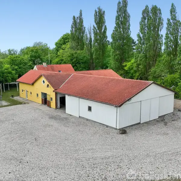 En gul bygning med rødt tegltag står blandt træer, omgivet af en stor grusbelagt gårdsplads. Til venstre ses en indhegnet sportsbane. Træer i baggrunden omkranser området.