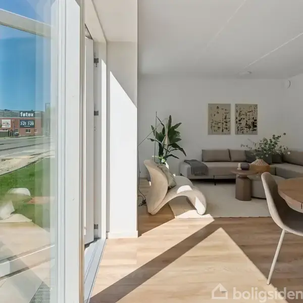 En åben terrassedør fører ind i en stue med lyse træplankegulve. En beige hjørnesofa står imod væggen ved siden af et sofabord. Udenfor ses en vej og en bygning med FedEx-skilt.