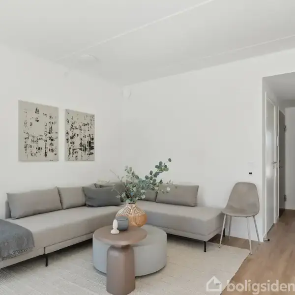 En grå sofa står i hjørnet af en stue med to små borde foran. Væggene er hvide, og et stykke kunst hænger over sofaen. En gang er synlig til højre.