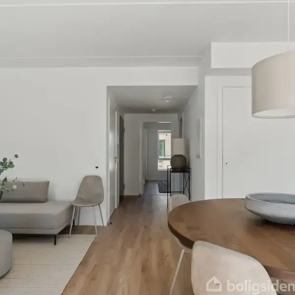 En moderne stue med minimalistisk indrettet sofa og bord med dekorative genstande, placeret mod en hvid væg. Til venstre et træbord med lampe. Lyse træplankegulve dominerer rummet.