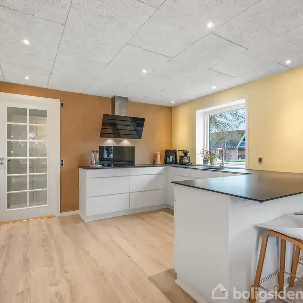 Køkken med hvidt køkkenø, to barstole og indbyggede hvide skabe. Rummet har en glasdør og trægulv. Et vindue giver udsigt til en have. På væggen findes en ugeplan.