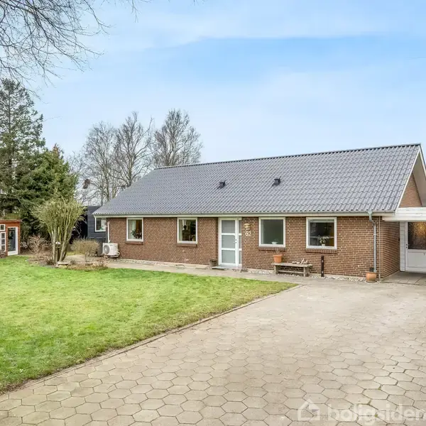 Et murstenshus med sort tag står foran en flisebelagt indkørsel. Til venstre er en velplejet græsplæne og et lille anneks i baggrunden. Træer omgiver området.