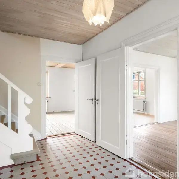 Entré med hvid trappe, der fører opad, flisegulv i røde og hvide nuancer. To åbne døre fører til lyse rum med trægulve, og en loftlampe hænger ned i midten.