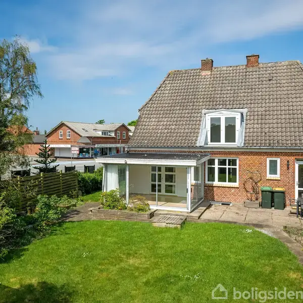 Et rødt murstenshus med gråt tegltag står stille. Foran er en grøn græsplæne omkranset af buske og træer. Nabohuse ses i baggrunden under en blå himmel.