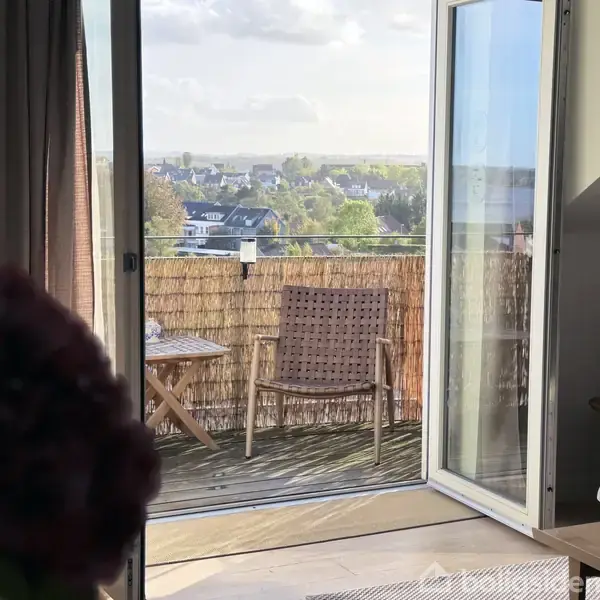 En balkon har en stol og et lille bord og ses gennem en glasdør. I baggrunden ses en byudsigt med huse og grønne træer.