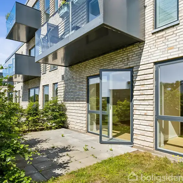 Flere moderne lejligheder med store vinduer og altaner strækker sig langs en bygning i lyse mursten, mens en lille terrasse og græsplæne fører ud til grønne buske under en blå himmel.
