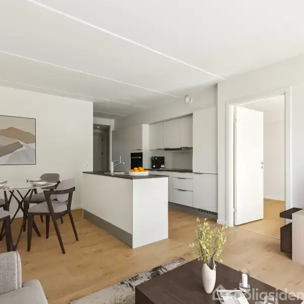 Stue med grå sofa, fremviser et fladskærms-TV på reol, spisebord med fire stole. Køkken med hvidt interiør, delvist synligt. Lyse trægulve og vægdekoration.