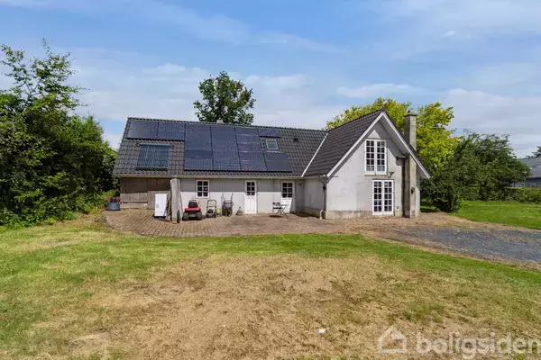 Huset står roligt i en grøn have med en stor græsplæne. Solceller er monteret på taget, og der er havemøbler på terrassen ved siden af huset.