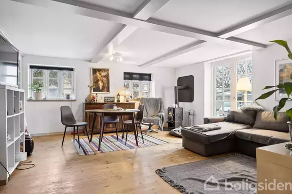 Stue med gråt sofaarrangement og brændeovn til højre. Rundt spisebord med stole til venstre. Trægulve, lyse vægge og synlige bjælker i loftet. Store vinduer lader lys ind.