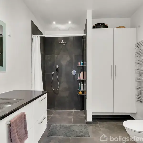 Et moderne badeværelse med en sort bruseniche har en sort væghængt bruser, hvide skabe, håndklædetørrer og grå fliser. En vask med spejl og sæbe beholder sig på venstre side.