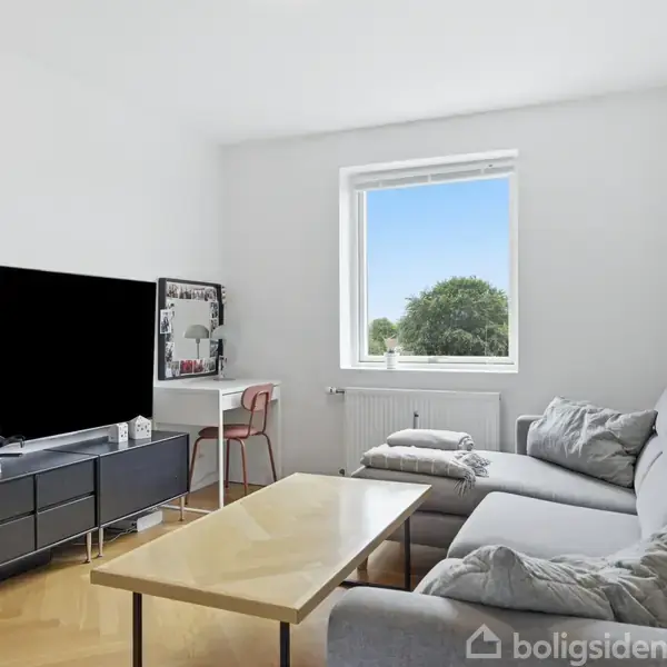 Grå sofa står i en lys stue med træbord foran. Et stort tv ses på et mørkt mediebord. Vinduet viser en klar himmel og træer udenfor.