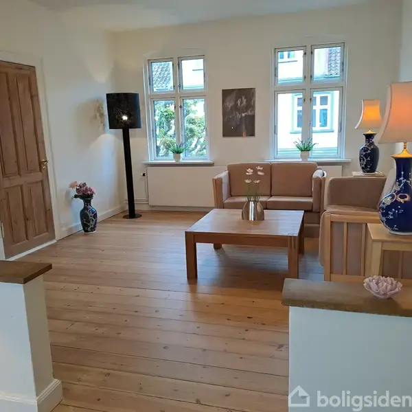 Stue med træmøbler og sofa, udsmykket med keramiklamper og planter. Vinduer giver udsigt til grønne omgivelser. Blomster og vægbilleder tilføjer dekoration til det lyse rum.