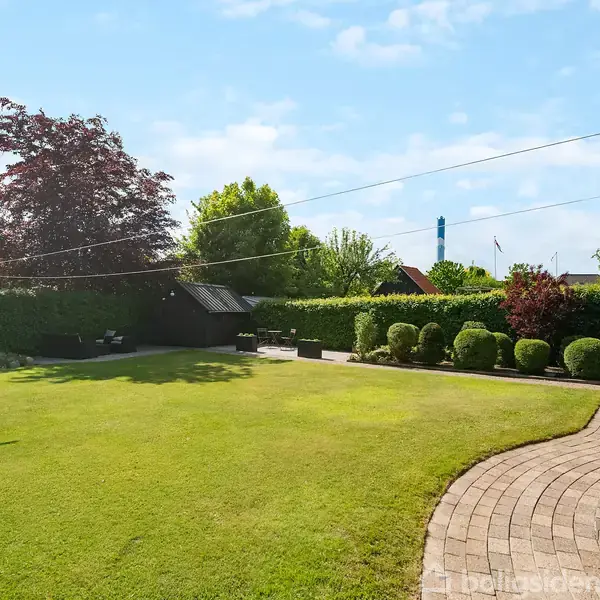 En velplejet have med en grøn græsplæne domineret af en træramme til tørresnor i en boligområde. En brolagt sti løber langs en huset facade mod en baggrund med buskadser og træer.
