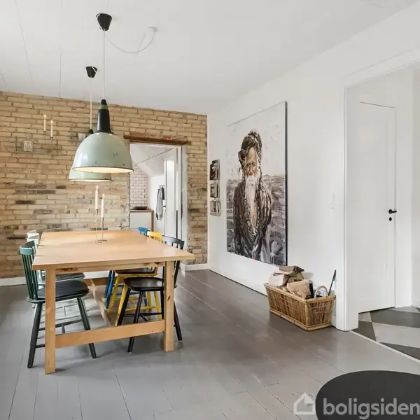 Et træbord med stole står i et moderne spisestueområde. En grøn hængelampe hænger over bordet. Brændeovn i hjørnet, murstensvæg til venstre. Billede af person på væggen.
