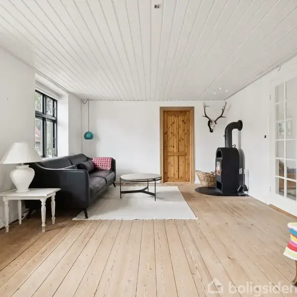 Sort sofa står mod en hvid væg i et lyst rum med trægulv. En brændeovn er til højre, nær en trædør og et hjortegevir på væggen. Et klaver er synligt ved glasdøre.