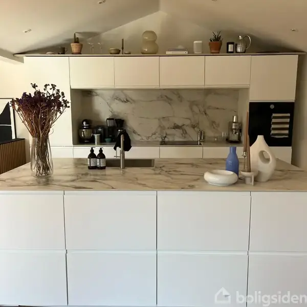 Køkken med hvid marmor bordplade, vase med tørrede blomster, og køkkenredskaber. Bagvæg i matchende marmor. Flasker og keramiske dekorationer tilføjer detaljer. Skabe og apparater i moderne stil.