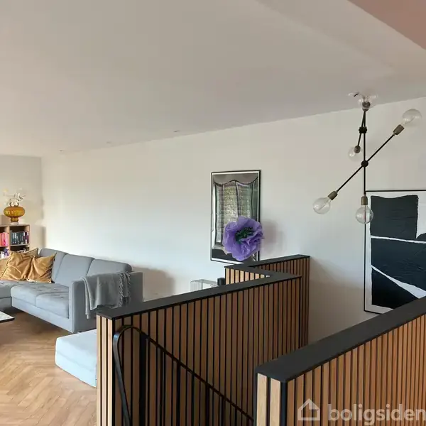 Grå sofa med puder står langs en hvid væg i en moderne stue med kunstværker og unik loftlampe. En dekorativ trappe er synlig i forgrunden.