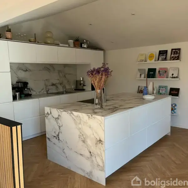 Køkkenø med hvid marmor, dekoreret med en vase med tørrede blomster. Indbyggede skabe og hylder med bøger i moderne køkkenindretning. En kaffemaskine står ved siden af vasken.
