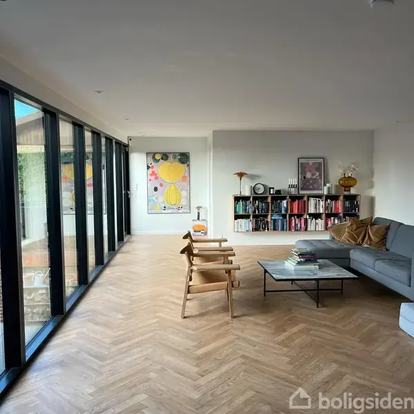 Stue med grå sofa, to træstole og et sofabord. Store glasvinduer til venstre giver udsigt til en terrasse. Reol med bøger og kunstværker hænger på væggen.