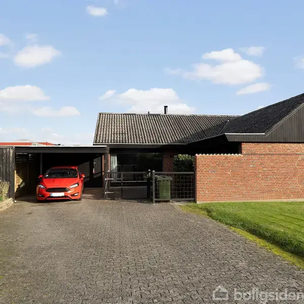 Rødt hus med fladt tag og carport, hvor en rød bil er parkeret. Omgivet af grøn græsplæne og hæk, under en blå himmel med spredte skyer.