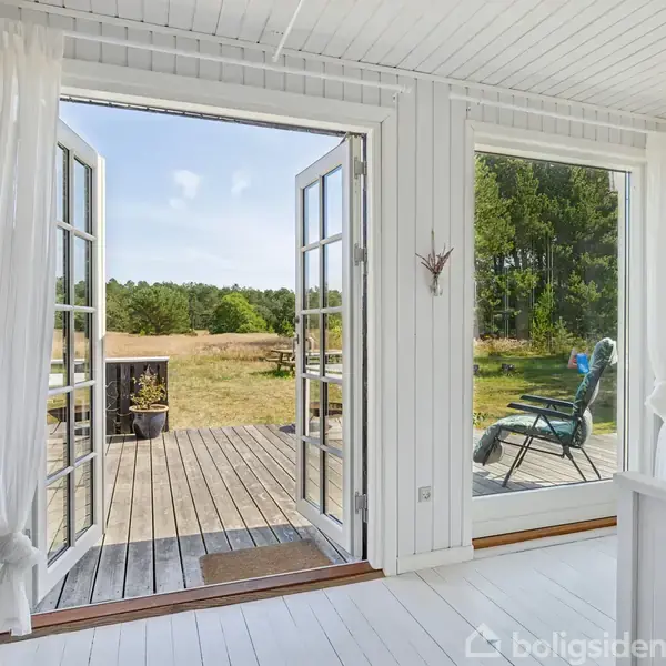 En hvidmalet stue med åbne franske døre fører ud til en træterrasse. I baggrunden ses en græsplæne og skov, mens en solstol står til venstre på terrassen.