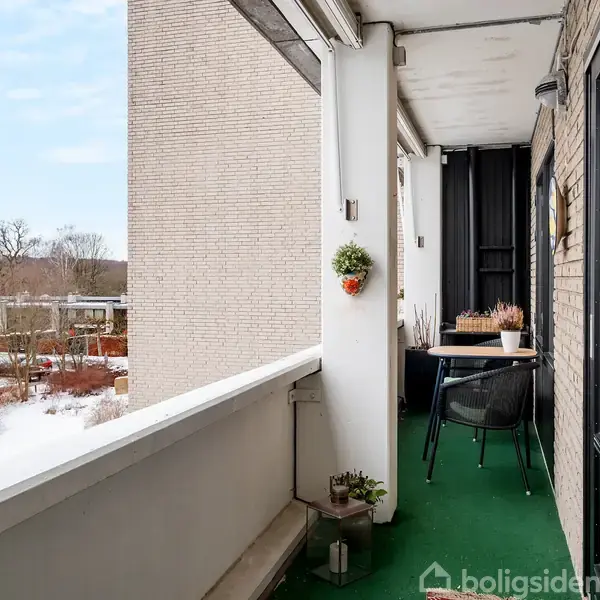 En altan med et lille bord og to stole står i et boligkompleks. Der er grønne planter på bordet og en hængende kurv på væggen. Udsigt til snedækket landskab.