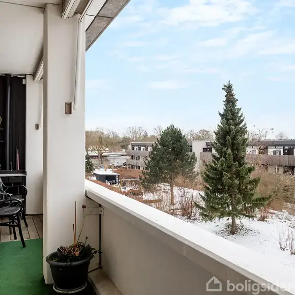 Altan med grønt tæppe og plante, to stole står ved bord. Udsigt til snebeklædt landskab med træer og rækkehuse under en klar blå himmel.