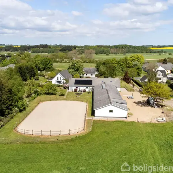 En gårdbygning med tilstødende ridebane omringet af grønne marker og træer i et landligt område. Flere huse ses i baggrunden under en skyet, blå himmel.