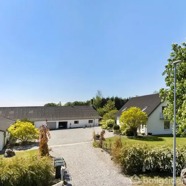 To hvide bygninger står på en grusvej omkranset af grønne træer og buske. Bygningerne er i landlige omgivelser under en blå himmel uden skyer.