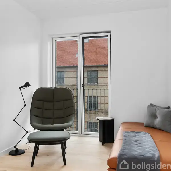 En moderne lænestol står ved siden af en gulvlampe i et lyst værelse med parketgulv. Ved siden af er en brun daybed med puder, og i baggrunden er et vindue med udsigt til en bygning.
