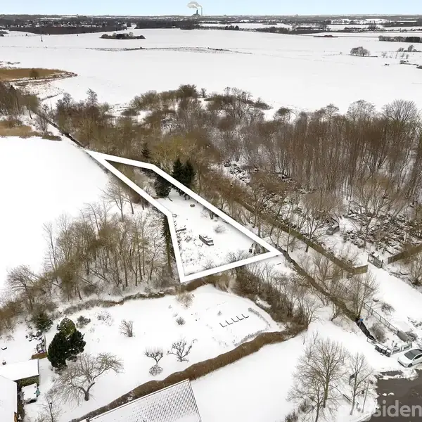 Et snedækket landskab ses ovenfra. En lang, trekantet grund er indrammet, omgivet af træer og marker. En vej med parkeret bil er til højre i billedet.
