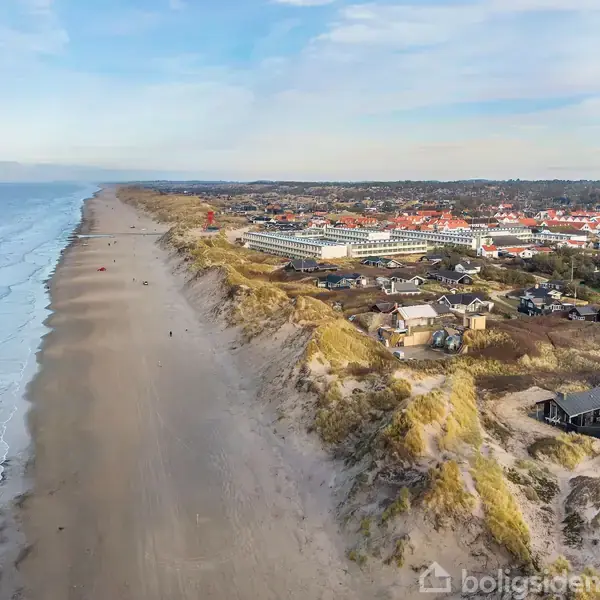 Strand strækker sig langs kysten med blidt bølgende vand på venstre side. Huse og bygninger ligger spredt til højre, omgivet af klitter, under en blå, let skyet himmel.