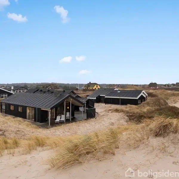 Et sort træsommerhus står stille mellem sandklitter og græs. Omkringliggende sommerhuse kan ses i baggrunden under en blå himmel med skyer.
