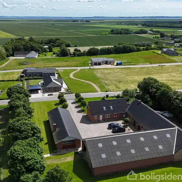 Luftfoto af en gård med tre bygninger i en U-form og geparkerede biler. Omgivet af grønne marker og spredte boliger i en landlig landskab.