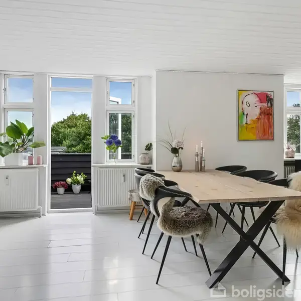Spisebord med sorte stole dækket af pels, står i et lyst rum med hvide vægge og trægulv. To store vinduer viser en udendørs altan med planter.