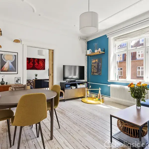 Et moderne køkken, hvor lange hvide skabe og en sort bordplade strækker sig langs væggen. En døråbning fører ud til en balkon med udsigt til en bygning med rødt tag.