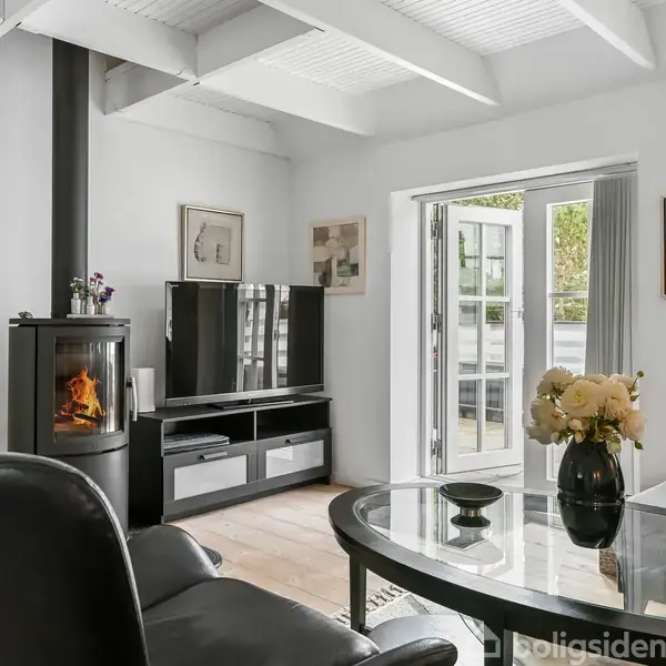 En stue med sorte læderstole omkring et glasbord med en blomstervase. En brændeovn brænder ved siden af et sort tv-bord. Store vinduer fører ud til en terrasse.