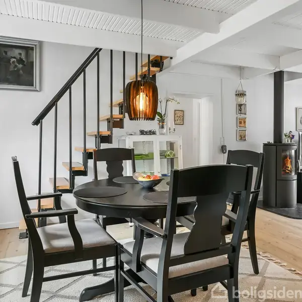 Et rundt spisebord omgivet af sorte stole står i midten af et lyst rum med trægulve. En trappe fører op ved siden af en reol. En kamin står mod væggen.