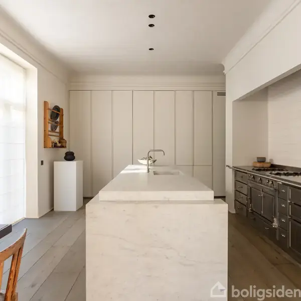 Et minimalistisk køkken med en central hvid marmor-ø og indbygget vask. Der er en lang, mørk bordplade til højre med komfur. Rummet har lyse vægge og trægulv.