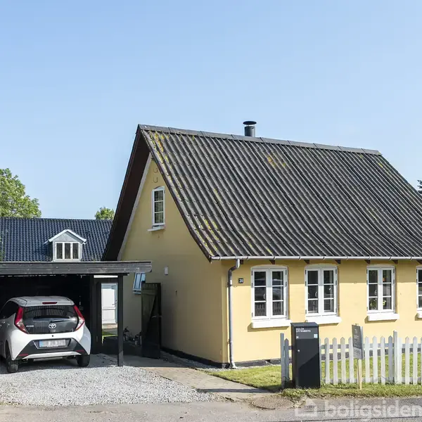 Et gult hus med sort tag, foran en hvid stakit. En bil er parkeret i en carport til venstre. Husnummeret "20" er synligt. Træer og flere huse omgiver huset.