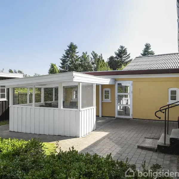 En gul bygning med en lille, hvid udestue, omgivet af en flisebelagt gårdhave og grønne buske. En sort dør til venstre fører til en anden bygning.