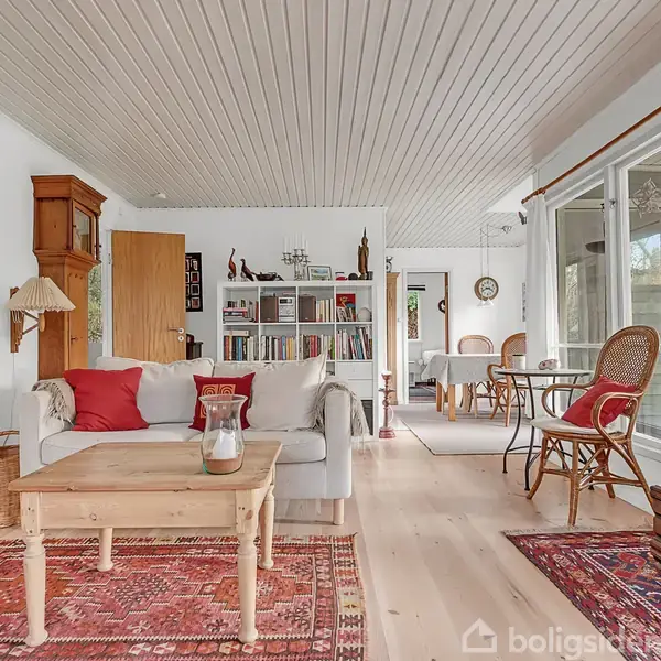 Stue med sofa og sofabord, dekoreret med farverige tæpper. Bogreol i baggrunden, store vinduer til højre, der giver udsigt til haven. Træmøbler og lys indretning skaber hyggelig atmosfære.