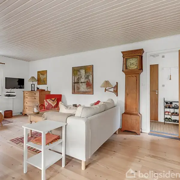 En stue med hvid sofa og træmøbler. Langvæg har bogreol og pendulur. Tv'et hænger ved vinduet med udsigt til have og blåt anneks. Malerier pryder væggen.