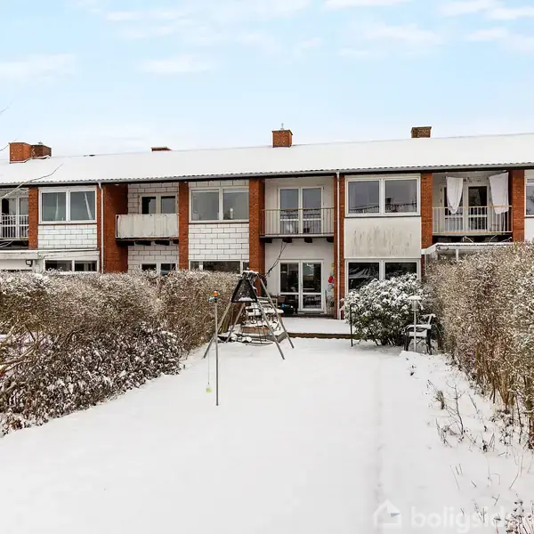 Et rækkehus dækket af sne, står med en snedækket have i forgrunden, omgivet af hække. En gynge er placeret midt i haven under en klar, blå himmel.