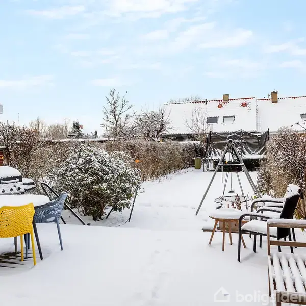 Terrassemøbler dækket af sne står i en snedækket have med en gyngestativ i baggrunden. Murstensvæggen til højre og buskene skaber en hyggelig, vinterlig atmosfære under en klar himmel.