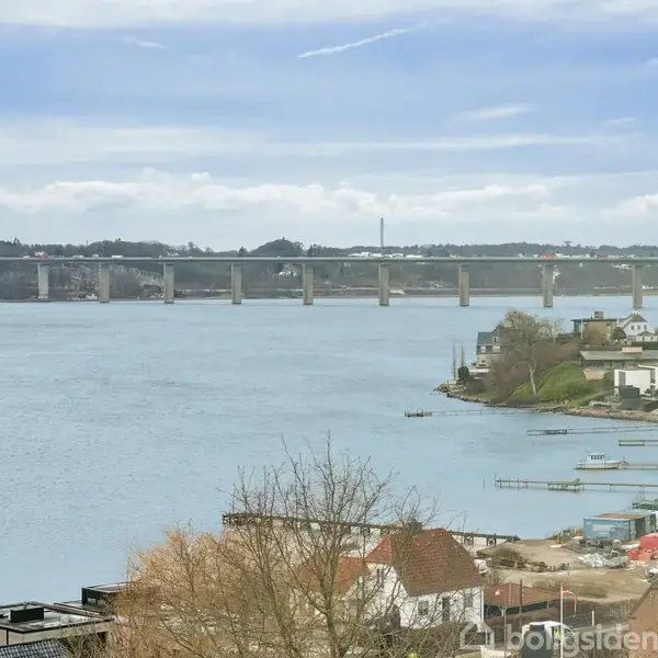 En bro strækker sig over en bred flod i et landskab med bakker i baggrunden. Forgrunden viser huse og træer langs vandkanten, der skaber en fredelig kystscene.