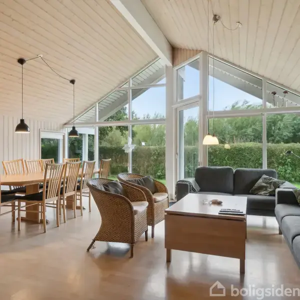 Stue med sofagruppe og træbord i forgrunden. Store vinduer bagved slipper naturligt lys ind, mens spisebord med stole står ved siden af. Trævægge og -loft skaber et hyggeligt miljø.