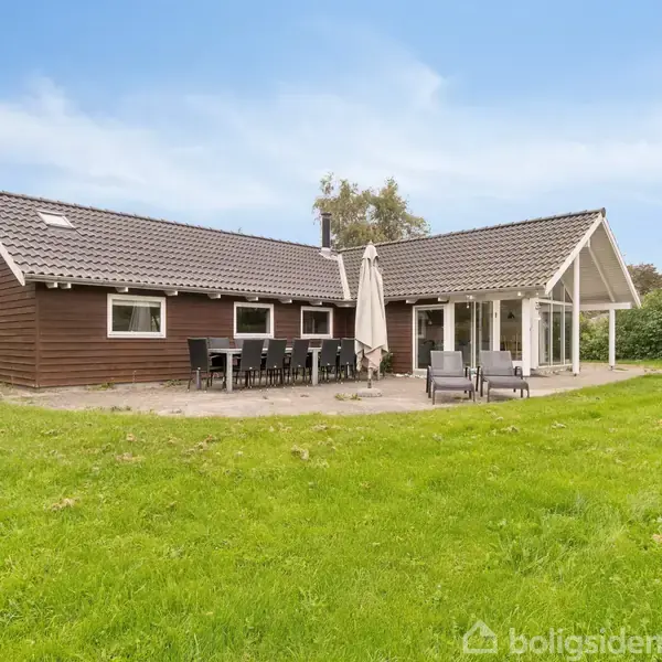 Et træhus står stille på en grøn græsplæne. Foran huset er der en terrasse med havemøbler og en stor parasol. Baggrund præges af træer og blå himmel.