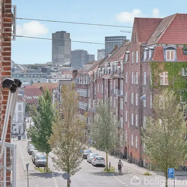 En byvej med murstensbygninger og træer på fortovet, omgivet af parkerede biler. Fjernt i baggrunden ses højhuse.